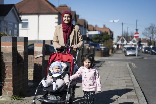 Front view of early 30s woman in hijab and warm clothing smiling at camera while enjoying exercise with toddler daughter and baby son in West London suburb.