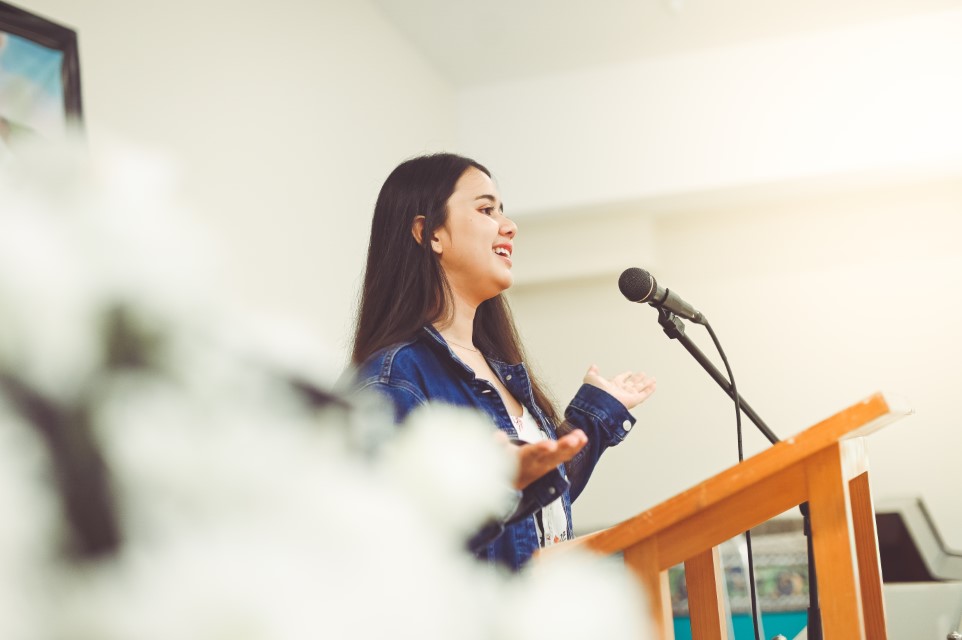 young lady, 18 years old, stands at a podium, maybe in school or at church and she gives a speech or perhaps she is singing. She is confident and accomplished looking. She is on the verge of something. She is mixed of, Asian, Thai and Latina ancestry