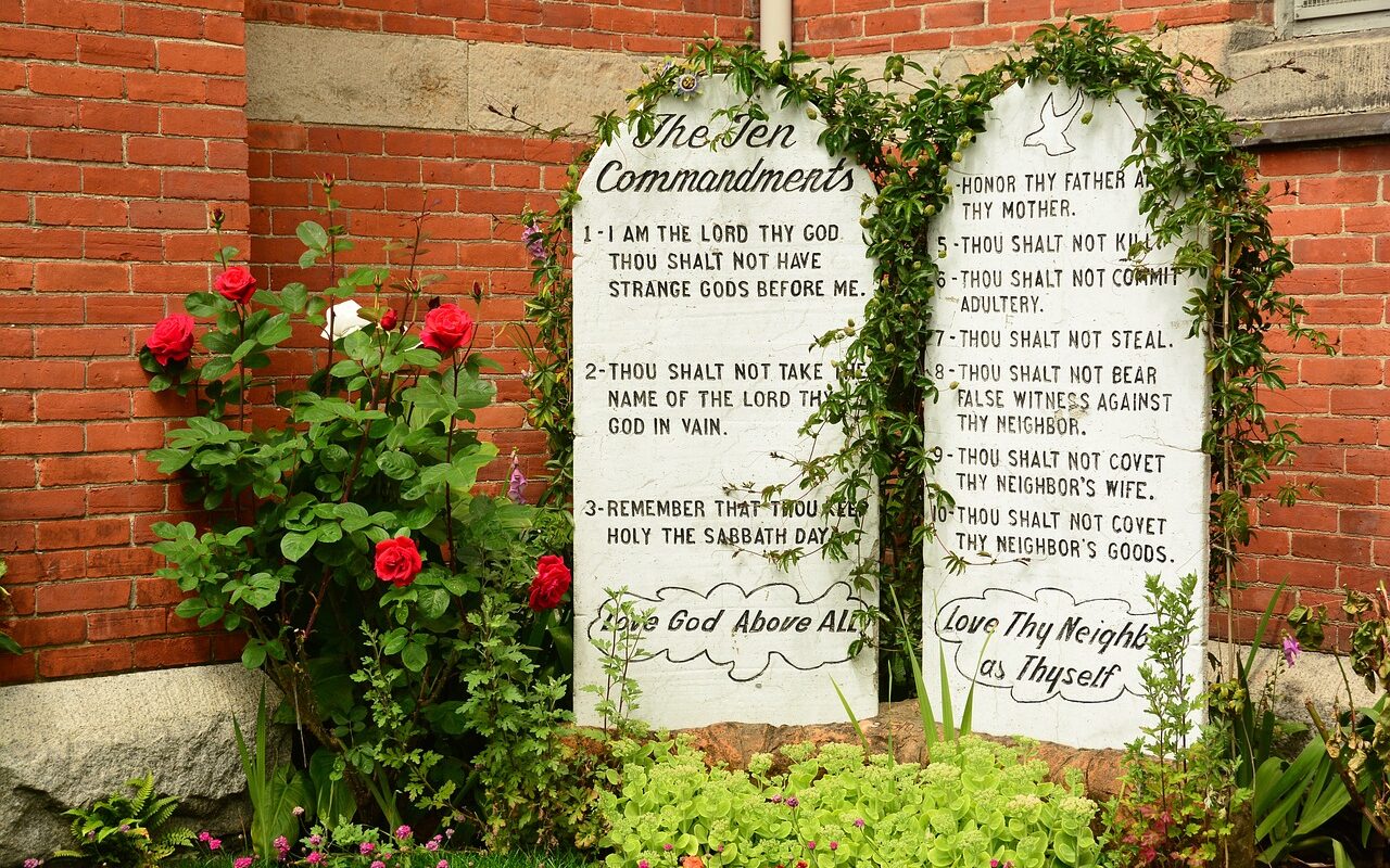 The ten commandments written on two stone tablets in front of a brick building and with green plants growing around the top edge of the stones.