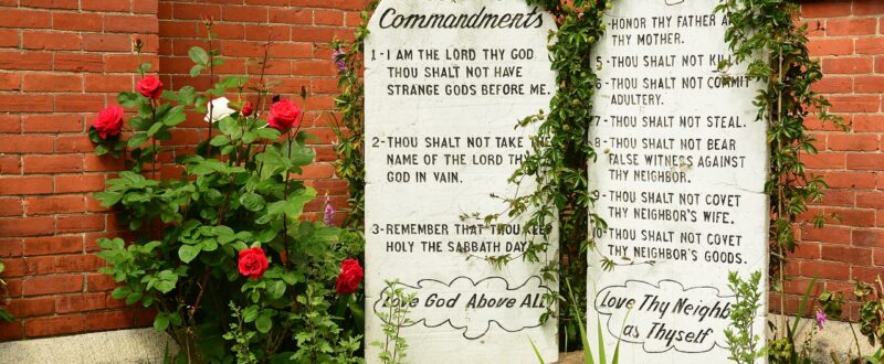 The ten commandments written on two stone tablets in front of a brick building and with green plants growing around the top edge of the stones.