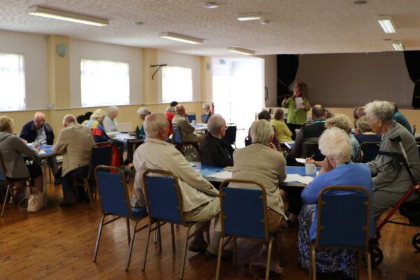 A group of people sat in a church hall facing away from the photographer listening to a speaker at the back of the image.