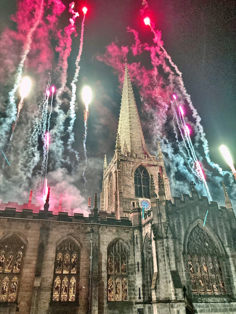 A photo of Sheffield Cathedral at night with multicoloured fireworks in the sky behind it.