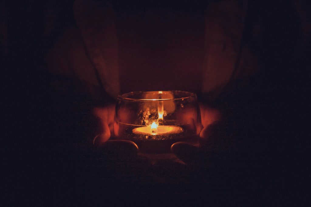 A pair of hands hold a tealight candle in the darkness.