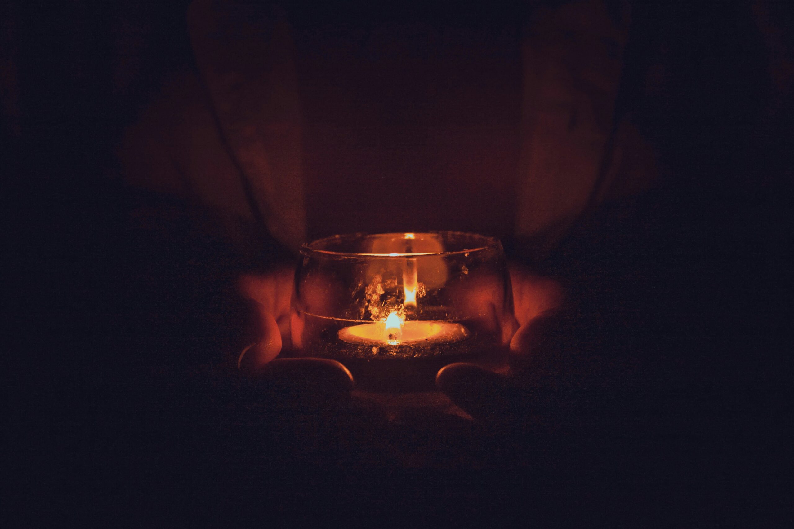 A pair of hands hold a tealight candle in the darkness.